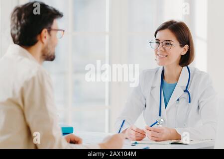 La foto di un paziente maschio scrive tutti i dettagli fa appunti durante la consulenza con il medico seduto in un ufficio spazioso. Il medico informa i problemi di trattamento e spiega Foto Stock