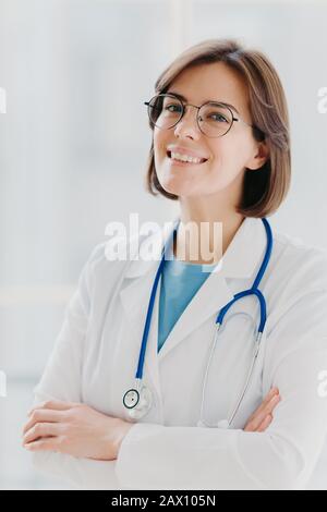 Primo piano ritratto di donna generale capelli corti si alza con sorriso e le braccia incrociate, usa stetoscopio, ama il lavoro, posa contro bianco b Foto Stock