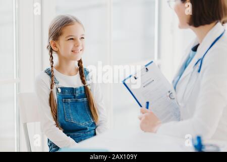 Felice bambino piccolo ascolta attentamente medici consigli e prescrizione, viene a vedere pediatra in clinica, ha due pigtail, indossa tute denim, t. Foto Stock