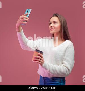 Una giovane bruna affascinante in un maglione e jeans tiene un caffè da andare e prende un selfie sul suo smartphone. Foto Stock