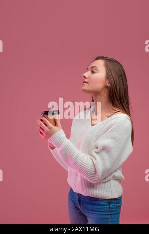 Affascinante giovane brunetta in un maglione e jeans tiene un vetro usa e getta per una bevanda calda, un ritratto verticale su uno sfondo rosa. Foto Stock