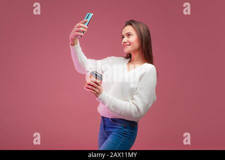 Una giovane bruna affascinante in un maglione e jeans tiene un caffè da andare e prende un selfie sul suo smartphone. Foto Stock