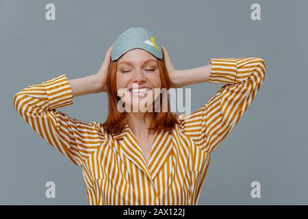 Concetto di pisolino diurno. Donna felice con i capelli zenzero indossa confortevole pigiama a righe e maschera di sonno, utilizza accessori utili per il sonno, sorrisi in generale, Foto Stock