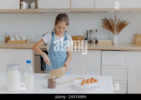 La ragazza carina indossa grembiule a righe, sussurra gli ingredienti nel recipiente, prepara l'impasto, insegna a cucinare, si alza al tavolo bianco con uova, latte, farina contro Foto Stock