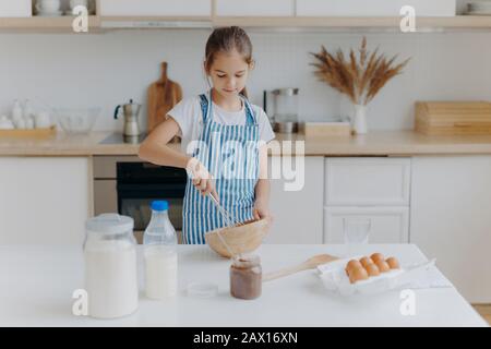 Piccola ragazza carina nel grembiule, mescola gli ingredienti, sussurra con frusta, usa uova, latte, farina, prova nuova ricetta, si leva contro l'interno della cucina, prepara t Foto Stock