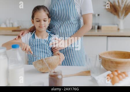 Immagine ritagliata di affettuosa madre in grembiule abbraccia la figlia che impara a fare pasta, tiene frusta, mescola gli ingredienti in ciotola, posa in cucina Foto Stock