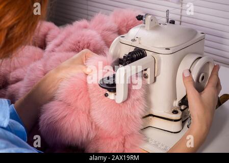 Master professionale per la produzione di capi di abbigliamento da pelliccia. Donna degno alla macchina da cucire cuce i vestiti da pelliccia rosa. Il processo di cucitura Foto Stock