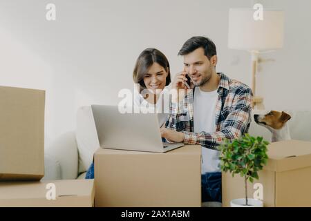 Un uomo felice dorme il laptop in un nuovo appartamento, chiama via smartphone, passa in un nuovo appartamento insieme a moglie, siediti su un comodo divano, circondato da un cardboa Foto Stock