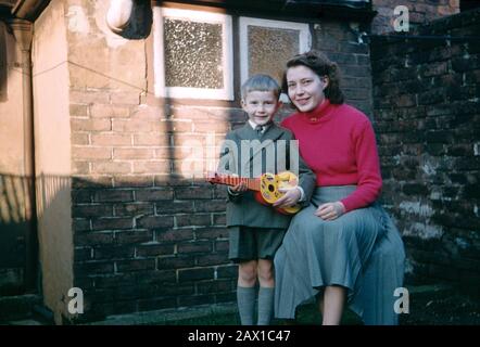 Ragazzo di cinque anni che indossa una tuta corta a due alberi e tiene una chitarra giocattolo che posa con sua madre nel cortile posteriore di una casa a schiera Foto Stock