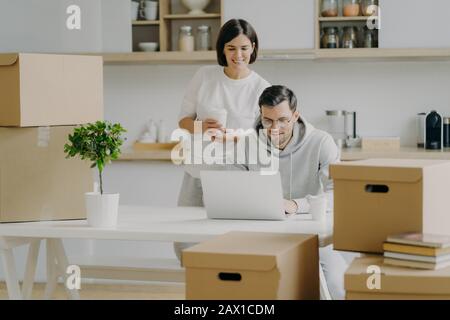 Foto di allegra giovane donna si trova dietro suo marito che lavora su computer portatile, posa in cucina moderna del loro nuovo appartamento, circondato da c. Foto Stock