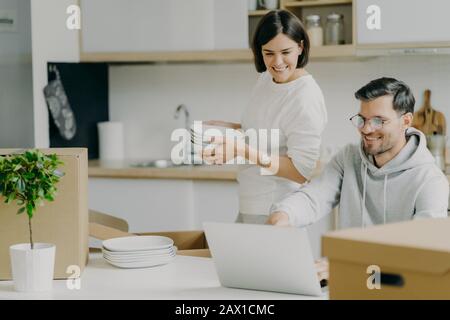 Casa in movimento e immobiliare. La donna bruna che si prende cura tiene un mucchio di piatti, scompatta gli effetti personali, il marito chiede il suo consiglio, sceglie qualcosa per b Foto Stock