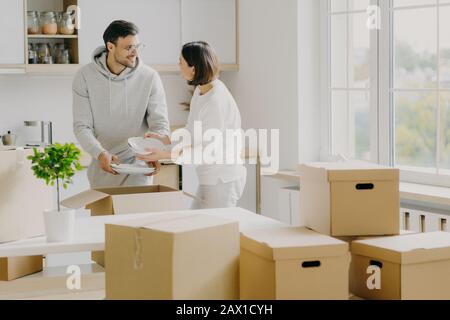 Foto di coppia occupato famiglia disimballare roba personale da scatole di cartone, vestiti in abiti casual, tenere piastre bianche, posa in spaziosa cucina con moder Foto Stock