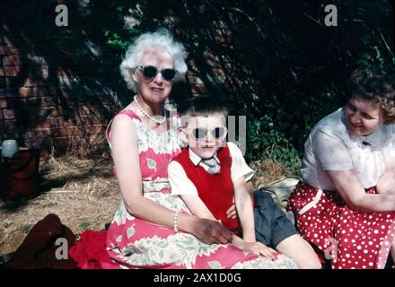 Ragazzo di sei anni che indossa occhiali scuri seduto con un pic-nic con i parenti più anziani nel 1959 Foto Stock