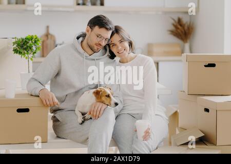 Stanco coppia sposata di famiglia bere caffè aromatico takeaway, sedersi a stretto contatto, posa con cane pedigree vicino a grandi scatole di cartone, posa contro kitc Foto Stock