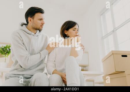 Foto di triste donna lamenta problemi, l'uomo calma, abbraccia moglie, stanco dopo aver portato molte scatole con le cose alla loro nuova casa, indossare un coagulo casual Foto Stock