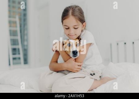 Indoor shot di felice piccolo bambino abbraccia preferito animale domestico, vestito in pigiami, esprime l'amore per il suo cane, gioca con animale dopo il risveglio, posa su così Foto Stock