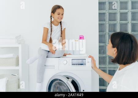 Adorabile bambina con pigtail, posa in cima alla lavatrice, tiene un panno morbido bianco, guarda gladfully alla madre, parla di piani dopo il lavaggio. Brunetta Foto Stock