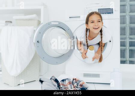 Felice ragazza europea con due trecce, dispone all'interno della lavatrice, contiene bottiglia bianca con polvere liquida, ha espressione premurosa, mucchio di clo sporco Foto Stock