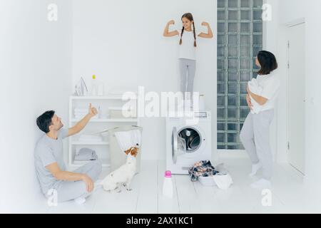 Immagine di felice bambino alza le braccia, mostra bicipite e stength, il padre mostra come segno con il pollice in su, stand in stanza di lavaggio con mucchio di vestiti in b Foto Stock