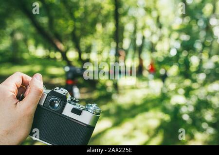 Una mano dell'uomo tiene una vecchia macchina fotografica vintage e fotografa un paesaggio verde. Vista in prima persona POV Foto Stock