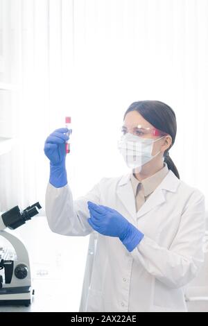 Ritratto di farmacologo femminile non riconoscibile che indossa camice da laboratorio, maschera protettiva. Guanti e tubo di prova per occhiali con liquido rosso Foto Stock