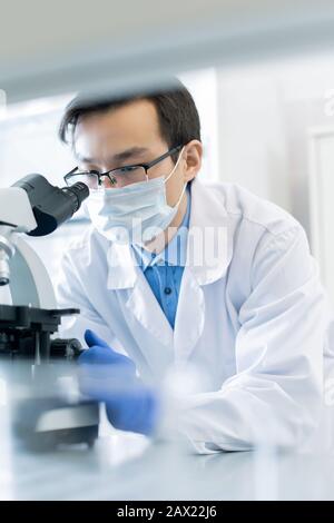 Ritratto verticale del giovane farmacologo asiatico che indossa maschera protettiva che esamina la reazione chimica dei campioni al microscopio in moderno laboratorio Foto Stock