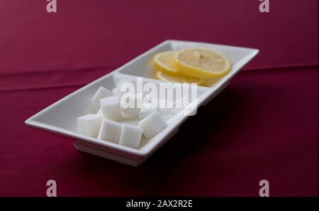 La selezione dei condimenti del tè ha i cubetti di zucchero dolci in prima linea del piatto bianco su stoffa rossa del tavolo Foto Stock