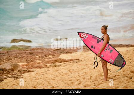 Surfista femminile che tiene surf rosa sulla spiaggia dopo aver cavalcato massicce onde in Banzai Pipeline sulla North Shore, isola di Oahu, Haleiwa, Hawaii, USA Foto Stock