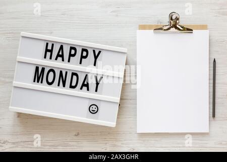 Parole 'happy monday' su una scatola luminosa, appunti con foglio di carta vuoto su una superficie di legno bianco, vista dall'alto. Sopra, dall'alto, disposizione piana. Primo piano. Foto Stock