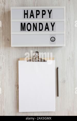 Parole 'happy monday' su una tavola moderna, appunti con foglio di carta bianco su una superficie di legno bianco, vista dall'alto. Sopra, dall'alto, disposizione piana. Chiudi Foto Stock