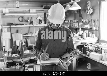 Ispettore all'interno del garage durante l'ispezione. Uomo caucasico di mezza età indossando casco e occhiali scrive note su un notebook, nero e bianco Foto Stock