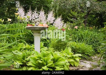 Sentiero in pietra e confine con bagno di uccelli, Astilbe rosa 'Venere', giallo, arancione e rosso Hemerocallis - fiori di giorno in giardino privato cortile Foto Stock