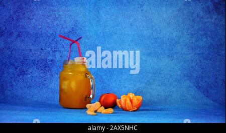 Una tazza di succo di mandarino con ghiaccio e cannucce accanto a pelati e tutta la frutta e fette di mandarino su uno sfondo blu. Close-up. Foto Stock