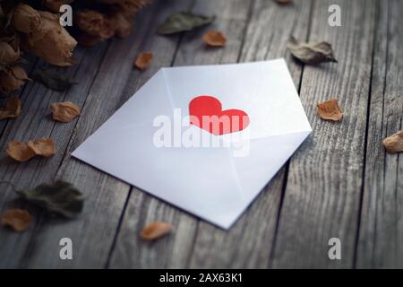 Busta aperta con singolo cuore di carta rossa incorniciato da rose secche su tavola di legno, confessione d'amore il giorno di San Valentino Foto Stock