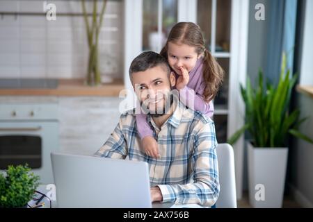 Padre seduto con un laptop, figlia che sussurra da dietro in orecchio. Foto Stock