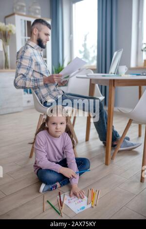 Scrittura figlia seduta sul pavimento, papà che lavora al tavolo. Foto Stock