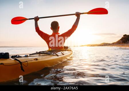 La vista posteriore dell'uomo tiene le pagaie del kayak alte. Attivo senior in kayak sul mare al tramonto Foto Stock