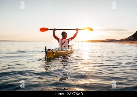 La vista posteriore dell'uomo tiene le pagaie del kayak alte. Attivo senior in kayak sul mare al tramonto Foto Stock