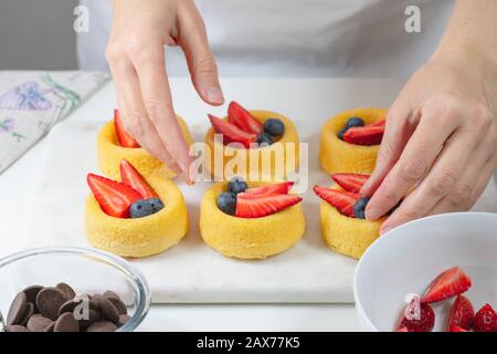Biscotti con frutti di bosco freschi, cioccolato e panna montata. Tazze fresche mini biscotto primo piano su sfondo bianco, mani donna Foto Stock