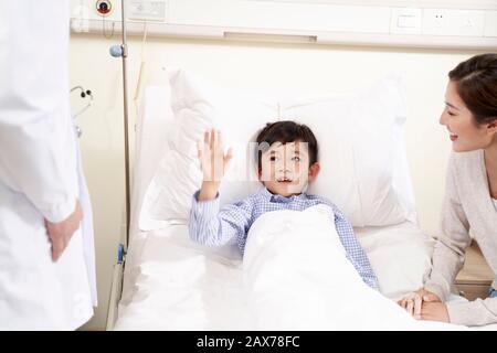 felice bambino asiatico di cinque anni sdraiato a letto nel reparto ospedaliero salutando il medico Foto Stock