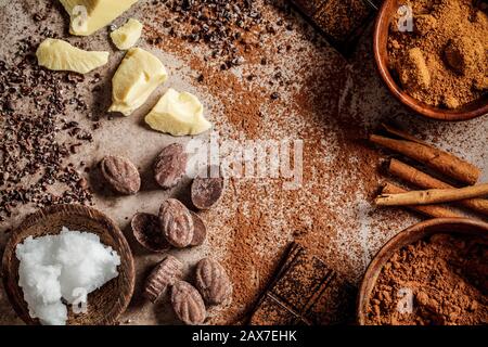 Ingredienti per la preparazione dello sfondo di cioccolato. Cacao, burro di cacao, zucchero, cannella e olio di cocco su sfondo scuro, copia spazio. Concetto di cioccolato. Foto Stock