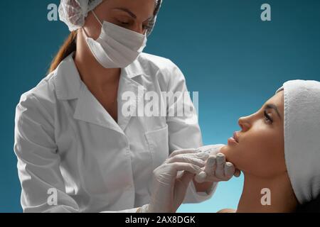 Vista laterale dell'iniezione nel mento femmina. Il cosmetologist in medici cappotto e maschera usando siringa con liquido speciale, bruna paziente seduta, isolato su blu. Concetto di cosmetologia, bellezza. Foto Stock