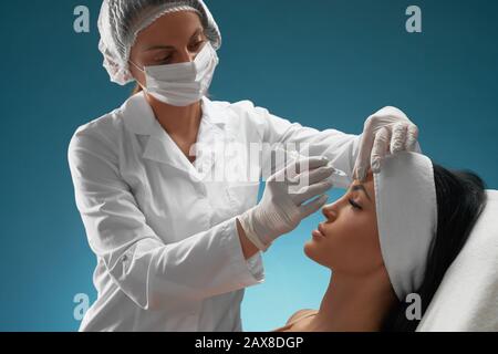 Vista laterale dell'iniezione nella fronte femminile. Il cosmetologist in medici cappotto e maschera usando siringa con liquido speciale, bruna paziente seduta, isolato su blu. Concetto di cosmetologia, bellezza. Foto Stock