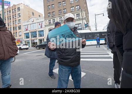 Un uomo americano cinese che indossa una maschera chirurgica porta fuori la lingua cinese che fa pubblicità a volantini su Main St. & Roosevelt Ave. A Chinatown, Flushing, NYC. Foto Stock