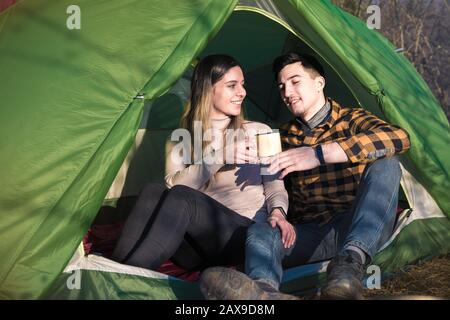 felice giovane coppia innamorata della natura sorridente, bere tè e caffè in tenda vicino a un fuoco. attività all'aperto per le famiglie giovani Foto Stock