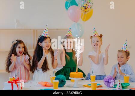 Gli amici dei bambini con gioia si aggrappano alle mani e si sguardi alla grande scintilla sulla torta, si alzano vicino al tavolo festivo con i presenti, le tazze e gli attributi di festa, hanno t divertente Foto Stock