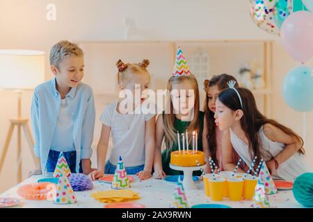 I bambini piccoli celebrano la festa di compleanno, soffiano le candele sulla torta, si riuniscono al tavolo festivo, hanno buon umore, godono trascorrere il tempo insieme, fare il desiderio, indossare la festa Foto Stock