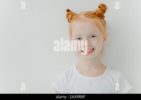Bambina positiva e attraente con doppio zenzero, fremiti sul viso, sorriso toothy, vestita con maglietta casual, si erge su sfondo bianco, e. Foto Stock