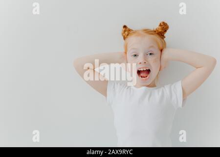 La ragazza redhead impressionabile tiene la bocca ampiamente aperta, copre le orecchie, eccitate dall'atmosfera rumorosa, indossa la t-shirt casual bianca, reagisce emozionalmente su qualche sottile Foto Stock