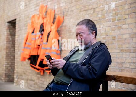 Berlino, Germania. 11th Feb, 2020. L'artista cinese ai Weiwei è seduto accanto al lavoro nella 'versione a parete' della sua opera 'Giacche di sicurezza Zipped l'Altro Modo' nel suo studio e guardando il suo smartphone. Le varie costruzioni di giacche di sicurezza, ganci e barre di ferro possono essere acquistate come parti individuali dallo sponsor Hornbach e assemblate da parti interessate stesse. Credit: Christoph Soeder/Dpa/Alamy Live News Foto Stock